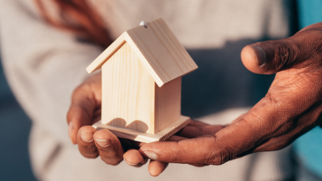 Hands gently holding a small wooden house model, symbolising housing, care, or support for a home.