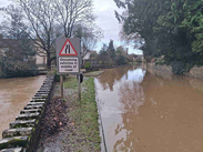 Flooded residential road with muddy water covering the carriageway, warning sign showing oncoming vehicles in middle of road, trees and houses nearby.