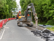 Excavator carrying out roadworks on a rural road, with temporary barriers in place as crews break up the road surface for repairs.
