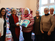 6 women standing in a row with blankets behind them. 2 women in the middle jointly holding a folded up blanket.