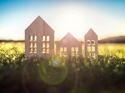 Three small wooden house models standing on grass in sunlight, symbolising homes, housing or communities in a natural outdoor setting.