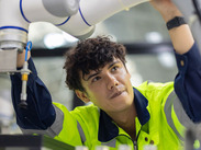 A young worker in a high-vis jacket operating a robotic arm in an industrial setting.