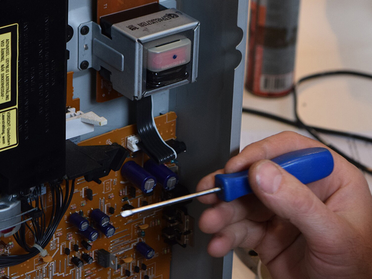 A hand holding a screwdriver repairing a opened DVD player.