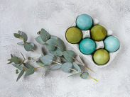 Green and turquoise Easter eggs arranged in a carton with eucalyptus leaves on a light, textured background.