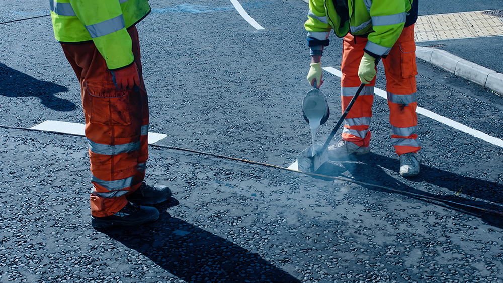 Roadworker applying thermoplastic road marking on the freshly laid tarmac during new roundabout and access road construction.
