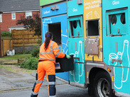 Recycling crew member in high-visibility gear loads sorted materials into a colourful recycling truck on a residential street.