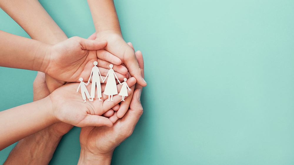 Hands of adults and children gently holding a paper cut-out family against a teal background.