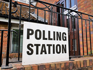 Polling station sign displayed on a gate outside a brick building, indicating a local polling place.