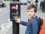 Child standing at a toucan crossing, pressing the pedestrian button beside the signal, with traffic and pedestrians in the background.