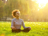 Happy young woman sitting in yoga position in a sunlit park, meditating on green grass surrounded by trees.