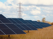 Solar panels installed in a rural field, with electricity pylons and overhead power lines visible against a blue sky with clouds.