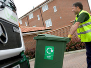 A worker in a reflective vest pushes a green garden waste bin towards a recycling truck in front of residential buildings.