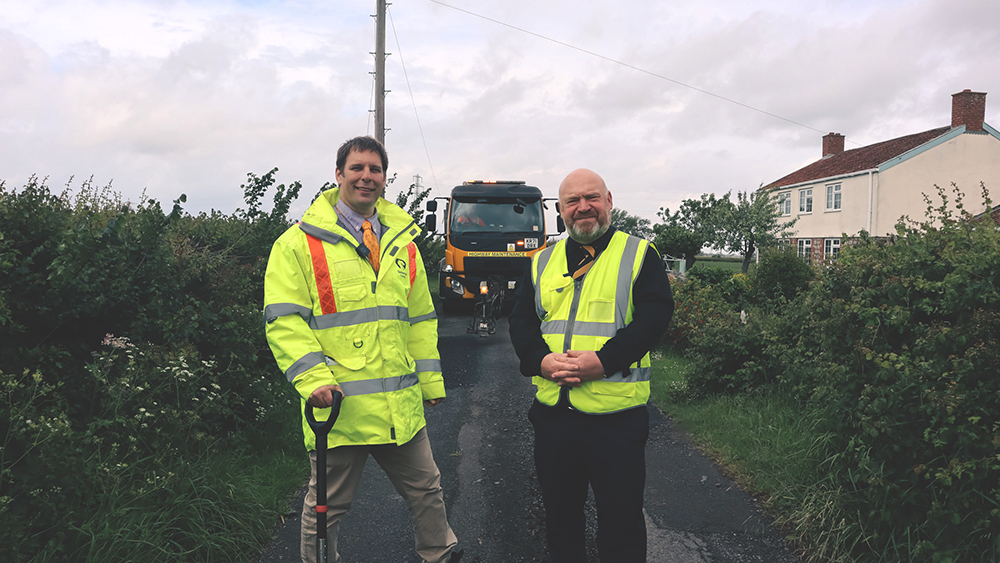 Cllr Wilkinson (left) and Cllr Revans stand on A30 Hendford Hill with highways vehicle behind during repair works.
