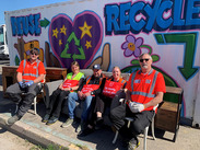 Recycling crew sit on donated furniture with reuse message mural behind, promoting bulky waste reuse through British Heart Foundation partnership.