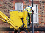 Worker in high-visibility clothing uses a raised platform to maintain or install equipment on a street pole outside a brick building.