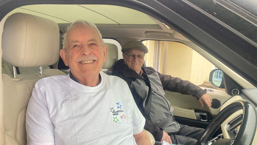 A senior volunteer driver wearing a Volunteers’ Week T‑shirt smiling from the seat of his car whilst transporting a passenger.