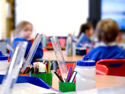 Classroom scene with focus on rulers, pencils, and water bottles on a table; blurred background shows children in blue uniforms.
