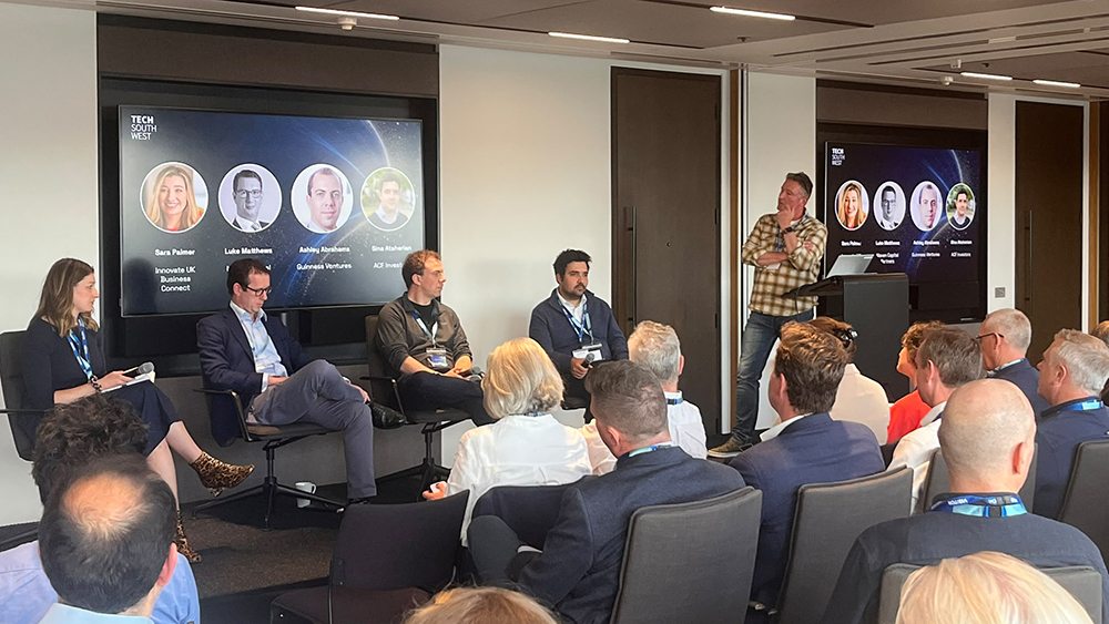 Panel discussion at a business event, with speakers on stage and audience listening, screens showing investor profiles and tech sector topics.