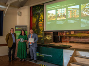 Cllr Smith-Roberts (centre) and Cllr Revans (right) receive CPRE Somerset award on stage with housing project presentation in background.