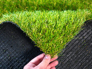 Hand holding a rolled section of artificial grass, showing green blades on top and black backing underneath.