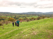 Walkers climb a grassy hillside at Dolebury Warren in the Mendip Hills, with patchwork fields and cloudy sky. Credit: Lucy Masters
