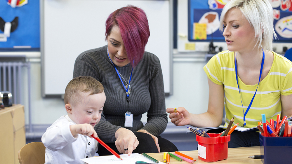 Adult and young child drawing together at a table with support from two staff in a classroom setting