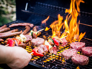 Burgers, sausages and vegetable skewers cooking on a barbecue grill with flames, as a hand turns the food.