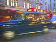Motion-blurred hackney carriage travelling through a city street, with buses and illuminated buildings in the background.