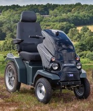 A black all-terrain mobility scooter parked on grass with rolling countryside and trees in the background.