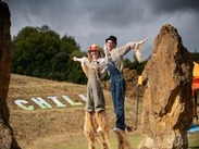 2 stilt-walkers dressed as scarecrows pose by standing stones at Ham Hill, with large white letters spelling “CHILL” on the hillside behind them