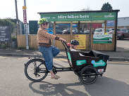 Person riding a cargo bike with a child passenger outside a Frome community bike hire hub, promoting car-free travel.