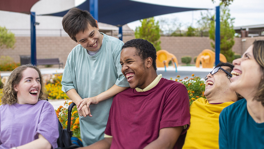 People laugh with their care worker in an outdoor community setting, sitting together and enjoying time in a garden space.