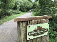 Sign for The Strawberry Line Path beside a tree-lined walking and cycling route, with a cyclist travelling in the distance.