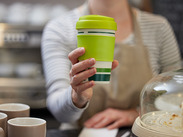 Café worker holding a green reusable takeaway cup across a counter, promoting sustainable alternatives to single-use drinks cups.