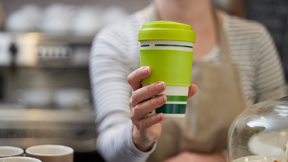 Café worker holding a green reusable takeaway cup across a counter, promoting sustainable alternatives to single-use drinks cups.