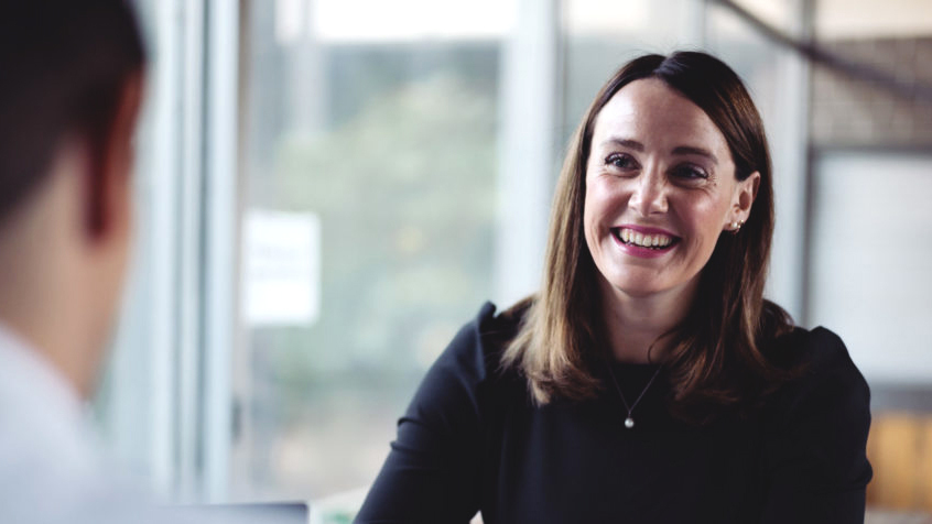 Woman smiling at a job interview across a table in a bright office, speaking with an interviewer in a professional setting.