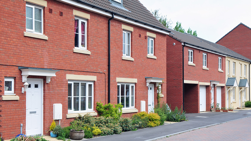 Modern red-brick terraced homes with front gardens, driveways and garages on a newly built residential street.