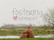 A man hugging a young person in a bear hoodie beside a river, with the Fostering in Somerset logo above.