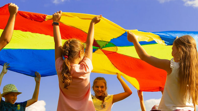 Children holding a colourful parachute overhead during an outdoor group activity on a sunny day.