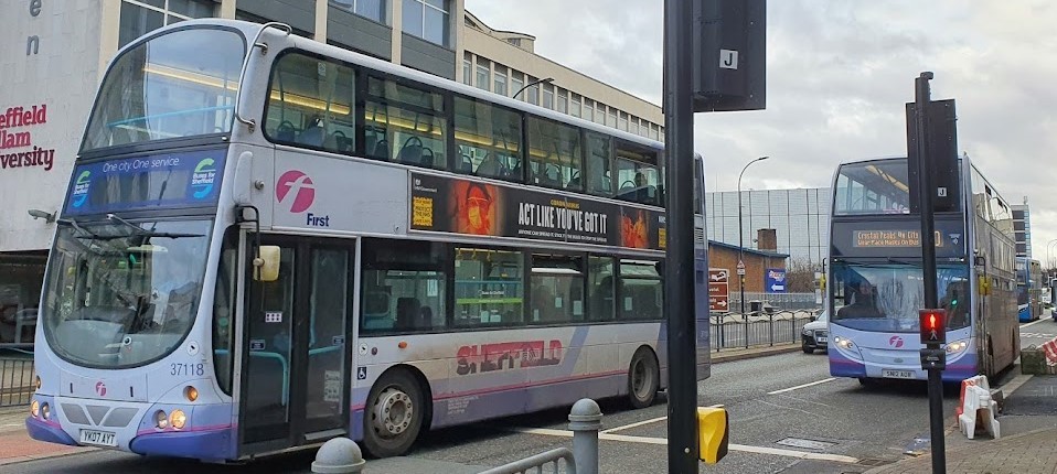 A sheffield city centre street, a double decker is driving past on the right followed by a single decker bus just behind.