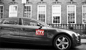 A black and white image of a taxi driving past a building. The City Taxi is in red on the door.
