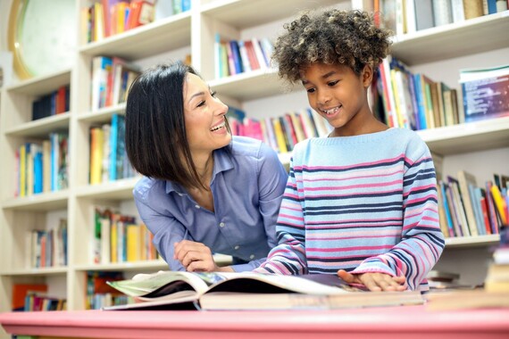 Smiling volunteer listens to a child reading in a classroom while sitting at desk