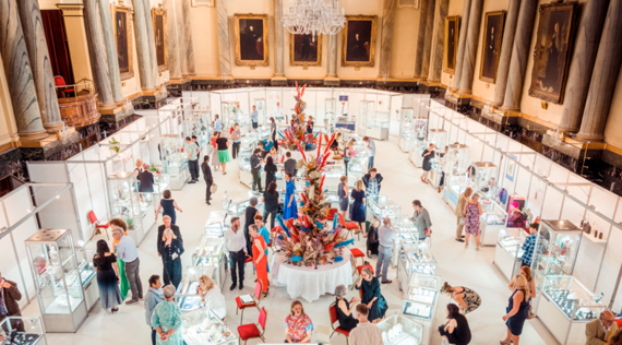 Jewellery and silverware displays in a grand hall with giant flower installations in the middle and people walking around.