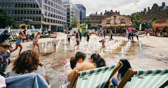 Children splashing in the fountains at the Peace Gardens with some people sat on deck chairs