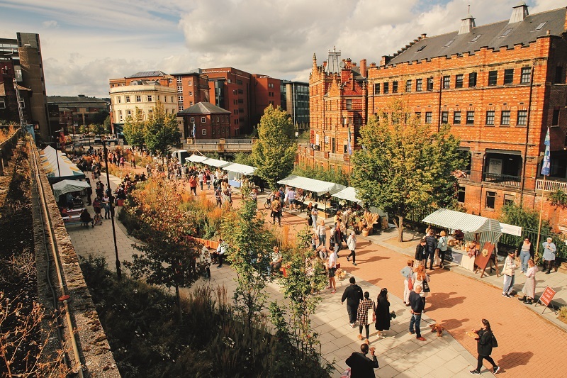 A crowd is gathered around the Pollen Market surrounded by old buildings and foliage.  