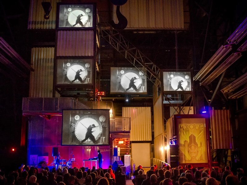 A crowd in a darkly theatre look up at a band with giant screens above their head displaying a person in a giant hamster wheel