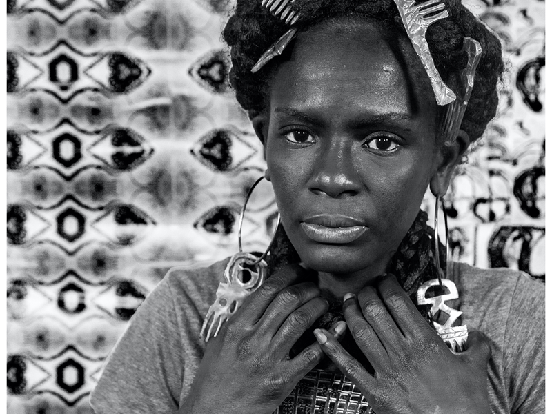 A black and white head-and-shoulders portrait of an adult with afro hair and their hands raised by their chin