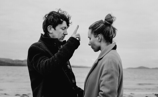 Image from the film called Communion. A windswept scene by the water a woman is being blessed by a man both bundled in winter coats.