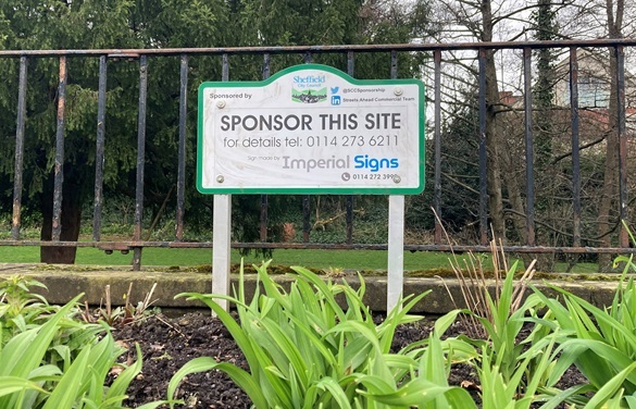 A blank sponsorship board stands amongst grass in front of a metal railing with trees in the background
