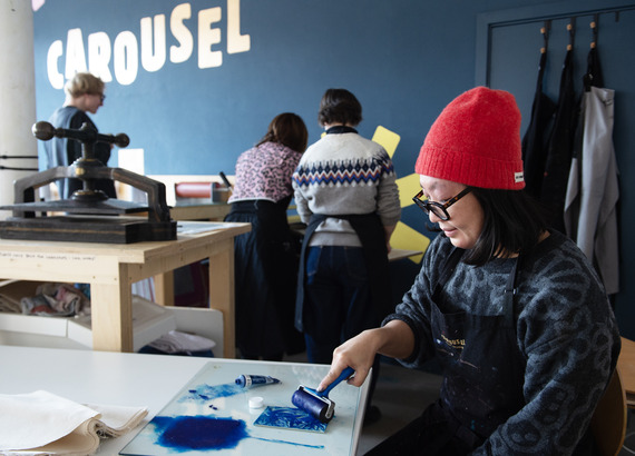 A person, wearing a red hat and an apron, is sitting and working on a printmaking project with blue ink.